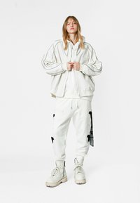 Sudadera blanca oversized con costuras en contraste, combinada con pantalones de chándal blancos que presentan detalles y parches negros. Botas de combate blancas completan el conjunto.