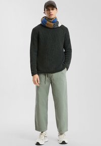 Pull en maille noir avec un motif texturé, pantalon large vert clair, et une écharpe épaisse multicolore, associés à des baskets blanches.