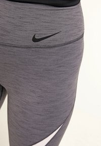 Szare legginsy sportowe wykonane z teksturowanej tkaniny, z czarnym logo Nike na pasku i białym akcentem po bokach.