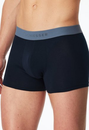 Männlicher Oberkörper, der dunkelblaue Boxershorts mit grauem Bündchen trägt, beschriftet mit "SCHIESSER", vor weißem Hintergrund.