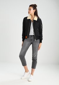 Giacca bomber nera con collo in pelliccia sintetica color crema, tasche laterali con zip, polsini a costine e una patch arancione, abbinata a jeans grigi cropped e sneakers bianche.
