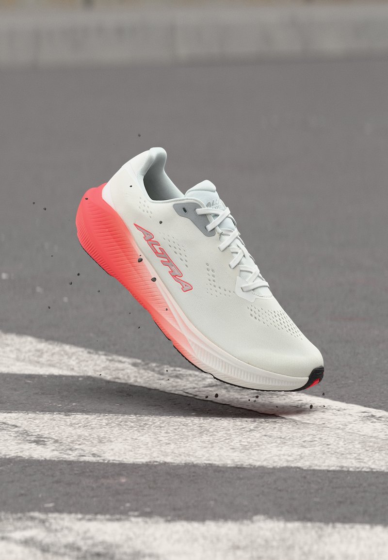 Λευκό και κοραλλί παπούτσι για τρέξιμο με κορδόνια, αιωρούμενο πάνω από διάβαση πεζών σε ασφαλτοστρωμένο δρόμο, με σωματίδια βρωμιάς γύρω του.