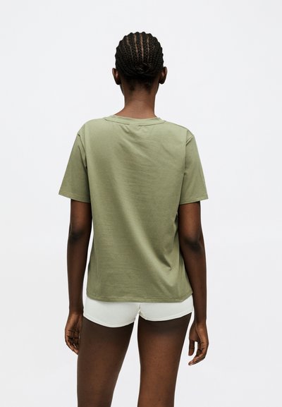 Personne aux cheveux tressés portant une chemise manches courtes vert olive et un short blanc, debout dos au spectateur sur un fond blanc.