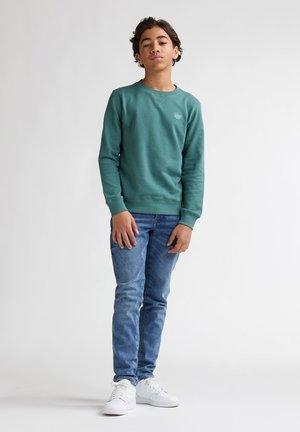 Felpa verde con polsini a coste, abbinata a jeans blu. Il modello indossa sneaker bianche e ha capelli ricci. Logo semplice sul petto.