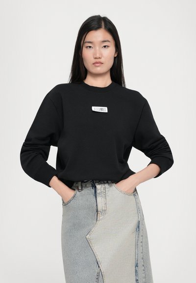Black sweatshirt with a round neckline and cuffed sleeves, featuring a small white label on the front, paired with a light blue denim skirt.