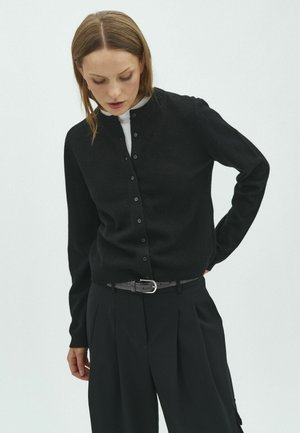 Femme portant un cardigan noir boutonné sur un haut blanc et un pantalon plissé noir avec une ceinture grise, regardant vers le bas avec une main sur la hanche.