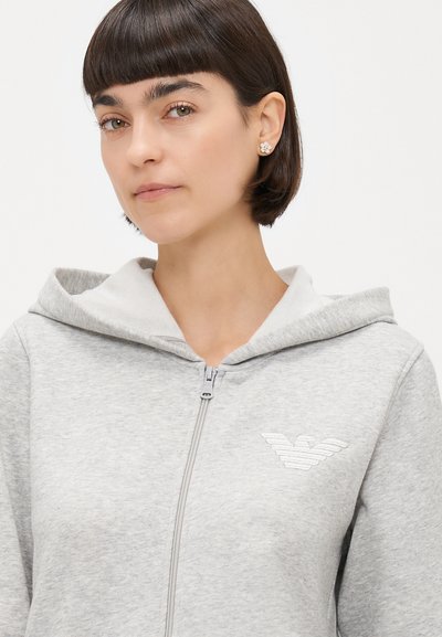 Pelēks zip-up džemperis ar mīkstu, teksturētu virsmu. Aprīkots ar odere kapuci un izšūtu logo uz krūtīm. Vienkāršs, minimālistisks dizains.