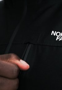 Χέρι που κρατάει το φερμουάρ ενός μαύρου μπουφάν North Face, έτοιμο να το κλείσει.