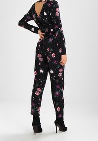 Czarny kombinezon w kwiaty z długimi rękawami, elastyczną talią i odkrytymi plecami. Charakteryzuje się różnorodnymi różowymi i białymi kwiatami na ciemnym tle.