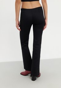 Czarne spodnie dzwony o gładkiej fakturze, w pasie średnim, z dwoma tylnymi kieszeniami. W połączeniu z czerwonymi botkami na obcasie, na neutralnym tle.