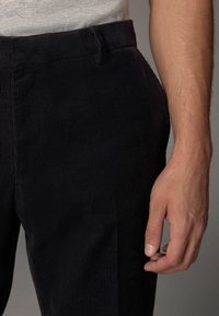 Schwarze Cord-Hosen mit feinenvertikalen Rippungen, die einen flachen Vorderbereich und einen Bund aufweisen. Der Stoff hat ein weiches, strukturiertes Aussehen.