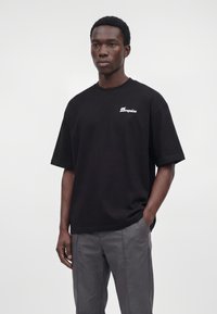 Jeune homme portant un T-shirt noir ample avec le texte "Dropsize" et un pantalon gris, debout avec une main dans la poche sur un fond uni.
