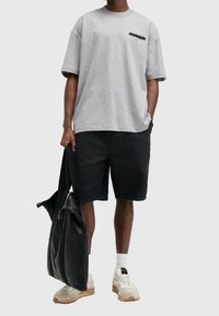 Camiseta gris de manga corta con un logo negro en el pecho, combinada con pantalones cortos negros, calcetines blancos y zapatillas beige. Bolsa de cuero negra.
