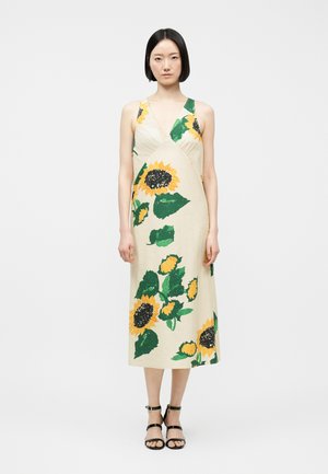 Žena v krémově bílé midi šatech bez rukávů s velkými potisky slunečnic a zelených listů, doplněná černými sandály na podpatku s pásky.