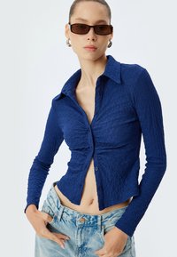 Camicia blu navy a coste con colletto, maniche lunghe e tessuto testurizzato. Abbinata a pantaloni corti in denim chiaro a vita alta.