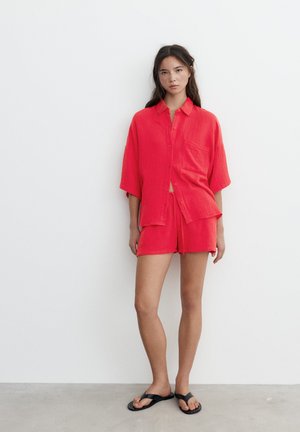 Femme debout contre un mur blanc portant une chemise rouge ample à boutons, un short assorti et des sandales tongs noires.