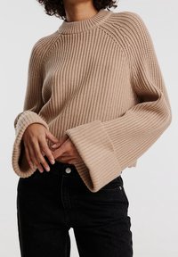 Suéter beige acanalado con cuello alto, mangas oversize y tela texturizada, combinado con jeans negros.