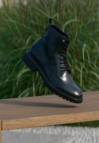 Stivale da combattimento in pelle nera con lacci sospeso sopra una panchina di legno con sfondo di erba verde all'aperto.