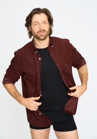 Mørkerød button-up skjorte med optrillede ærmer, båret over en sort T-shirt og mørke shorts. Glat tekstur, afslappet pasform og synlige knapper.