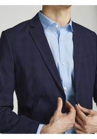 Marinblå blazer med subtilt rutmönster, enkel knappstängning, skräddarsydd passform, kombinerad med en ljusblå skjorta och kontrasterande knappar.