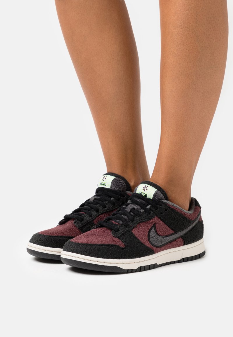 Person, der schwarze und burgunderfarbene Nike-Sneaker mit weißen Zwischensohlen trägt, steht vor einem weißen Hintergrund.