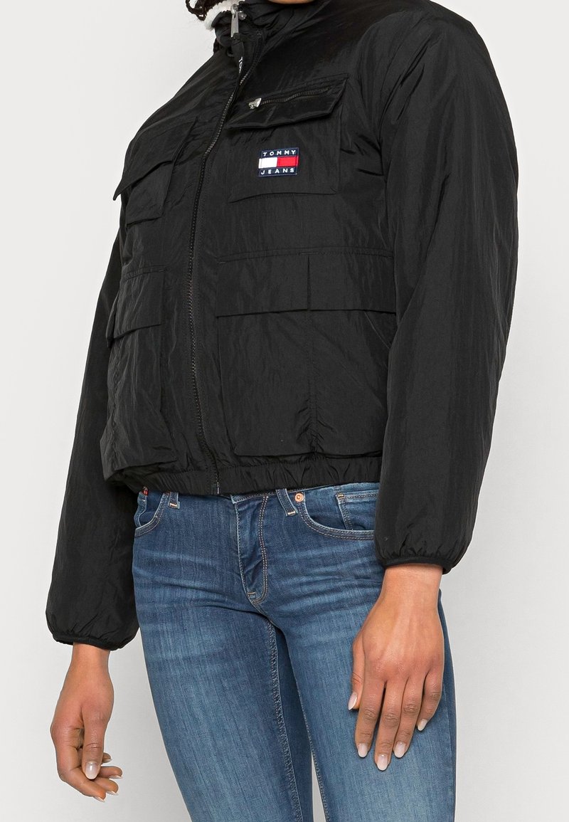 Czarna krótka kurtka z zapięciem na zamek, elastycznymi mankietami i wieloma przednimi kieszeniami. Posiada naszywkę z logo Tommy Jeans na górnej lewej piersi.