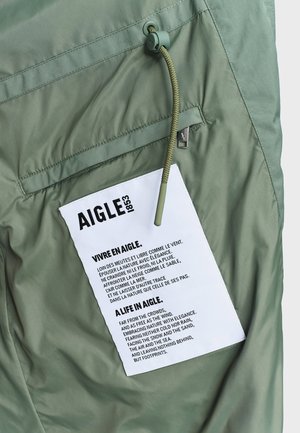 Light green jacket detail with zippered pocket and white fabric label featuring bilingual Aigle brand text and a green drawstring cord.