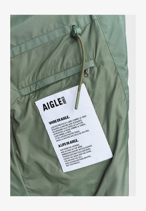 Light green jacket detail with zippered pocket and white fabric label featuring bilingual Aigle brand text and a green drawstring cord.