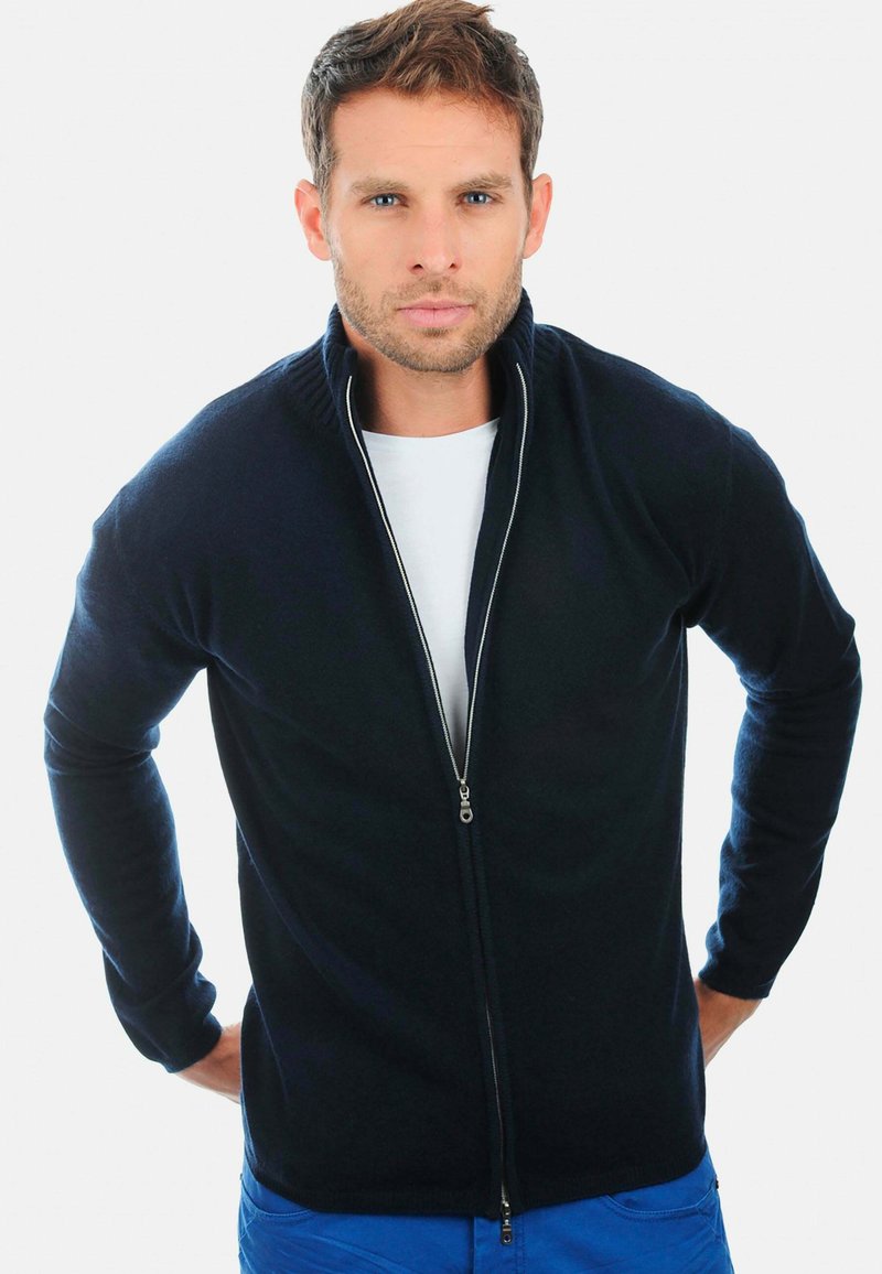 Homme avec des cheveux bruns courts portant un sweat à zip bleu marine sur une chemise blanche et un pantalon bleu, se tenant avec les mains derrière le dos.