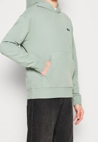 Sudadera con capucha verde claro hecha de forro polar, con un bolsillo frontal, puños y dobladillo acanalados; presenta un pequeño logotipo negro de la marca.
