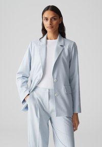 Blazer azzurro chiaro su misura in tessuto morbido, con rever a patta, doppi bottoni e tasche laterali, abbinato a pantaloni coordinati.