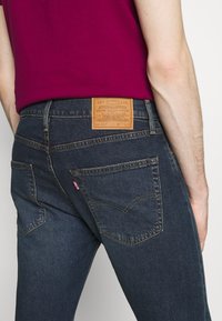 Donkerblauwe denim jeans met een leren patch met merkinformatie op de taille. Witte stiksels accentueren de achterzakken.
