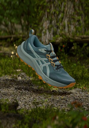 Chaussure de trail running bleue et orange avec semelle texturée suspendue au-dessus d'un sol forestier rocheux avec un fond de feuillage vert flou.