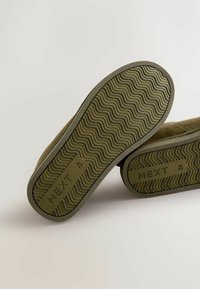 Olivengrøn fodtøj med teksturerede gummisåler, der har et bølget mønster. Mærket "NEXT" og størrelse "42" er præget på sålerne.
