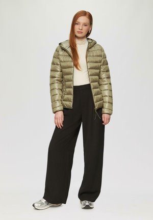 Olijfgroene, puffy jas met capuchon, uitgevoerd met een rits aan de voorkant en een verstelbare trekkoord. Gecombineerd met zwarte wijde broek en zilveren schoenen.