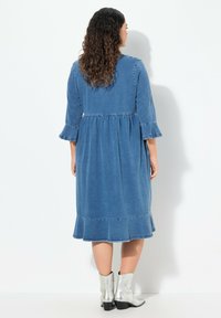 Kevätmekko farkkukankaasta, jossa on kolme neljännesosa hihoja, kerätty vyötärö ja röyhelöhelma. Ominaisuuksiin kuuluu vaaleansininen pesu, ja se on yhdistetty hopeisiin nilkkureihin.