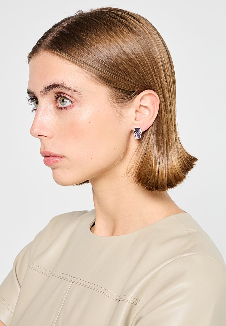 Młoda kobieta z krótkimi brązowymi włosami, ubrana w beżowy top i geometryczne fioletowe kolczyki, spoglądająca w lewo na tle jednolitego tła.