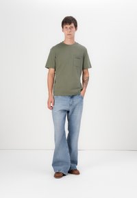 Olivenfarget bomull T-skjorte med brystlomme, sammen med lyseblå vide denimjeans. Modellen står mot en hvit bakgrunn.