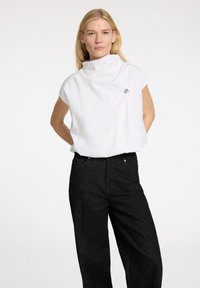 Camicia bianca a maniche corte con collo a imbuto e un piccolo accento metallico, abbinata a jeans in denim scuro, dal taglio morbido.