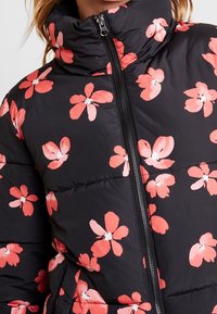 Svart pufferjacka med korallblommönster, med hög krage och frontdragkedja. Det quiltade mönstret ger dimension och värme.