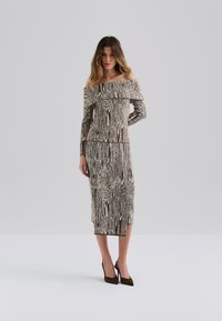 Abito off-shoulder con una trama testurizzata in un motivo legno marrone e crema, caratterizzato da maniche lunghe e una silhouette aderente, abbinato a tacchi scuri.