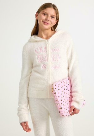 Weiße, flauschige Zip-Hoodie mit rosa gesticktem Text, kombiniert mit passender flauschiger Hose, die eine rosa Tasche mit floralem Muster hält.
