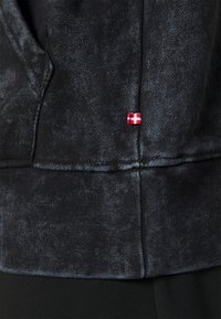 Černá mikina s texturovaným povrchem, žebrovaným lemem a malou nášivkou dánské vlajky v červené a bílé barvě poblíž spodního švu.