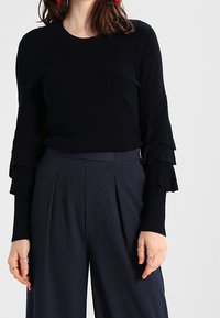 Black ribbed knit pullover with layered ruffle sleeves, paired with navy pinstriped high-waisted trousers. Accentuated with rings.