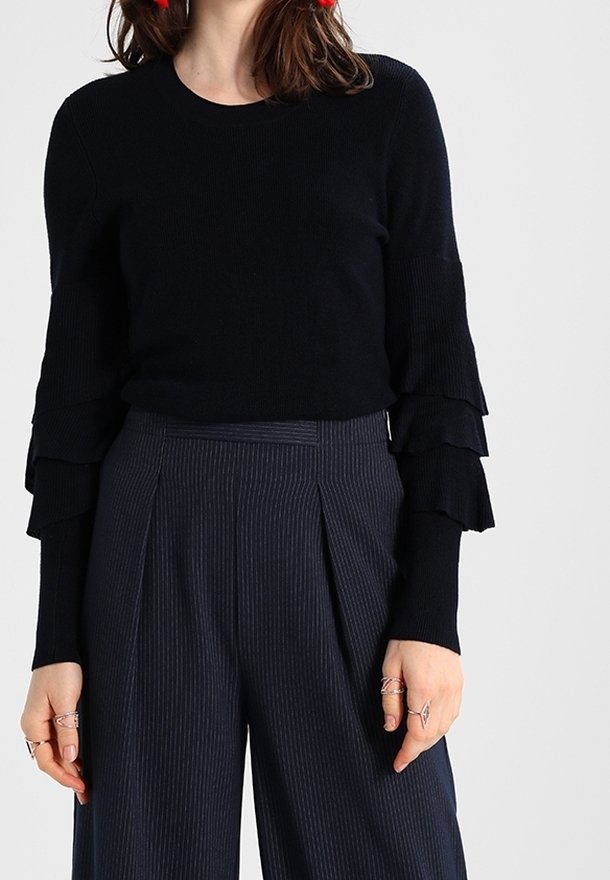 Black ribbed knit pullover with layered ruffle sleeves, paired with navy pinstriped high-waisted trousers. Accentuated with rings.