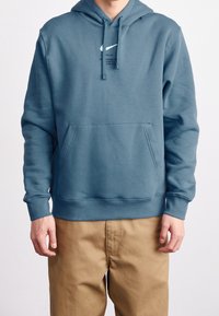 Nike blå hoodie gjord av mjukt tyg, med en känguruficka, huva med dragsko och vit logga på bröstet. Avslappnad passform och långa ärmar.