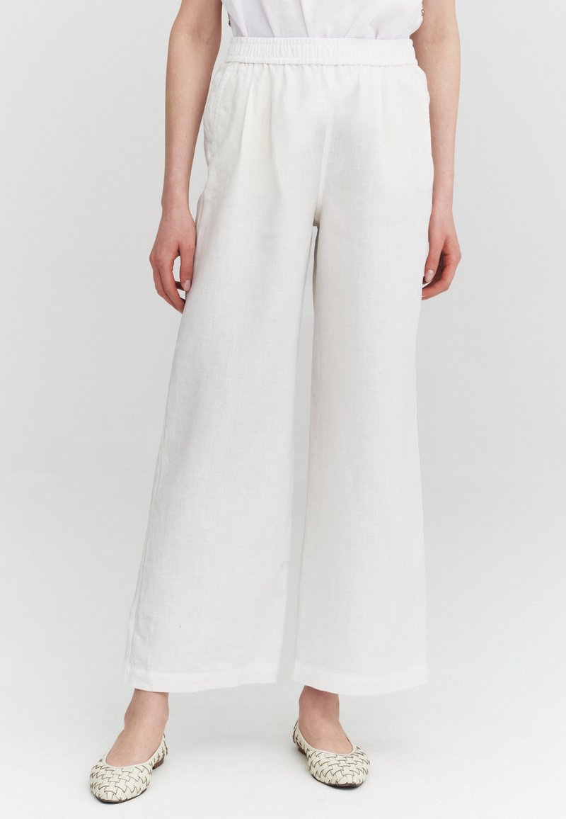 Femme portant un pantalon large blanc ample avec taille élastique et des ballerines beiges tissées, debout sur un fond uni.