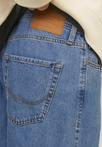 Zoom na zadnji žep modrih denim hlač s usnjenim logotipom blagovne znamke in rahlo dvignjeno bež vrhnjo oblačilo osebe.