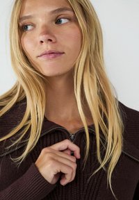 Burgunderfarbener Strickoberteil mit einem Reißverschlusskragen, der ein strukturiertes Muster und lange Ärmel hat. Haare locker über die Schultern drapiert.