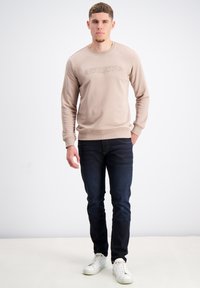 Felpa beige con scollatura rotonda e logo testurizzato "GABBJANO", abbinata a jeans blu scuro e scarpe da tennis bianche. Tessuto morbido, vestibilità casual.